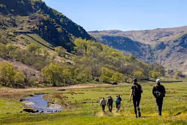 Photo of people walking across grassland besides a winding river with a back drop of hills.